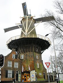 Photographie en couleurs d'un moulin à vent et de ses dépendances attenantes vus du dessous, le tout encadrés par un feu tricolore sur la gauche, un panneau signalétique et un arbre effeuillé sur la droite.