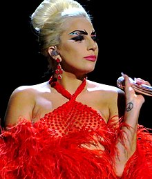  in a red feathery dress looking a little to her left, while holding a microphone in her left hand
