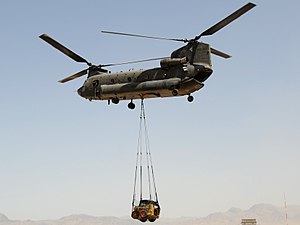 Colour photo of a green twin-rotored helicopter in flight with a yellow front loader suspended below the aircraft by a rope