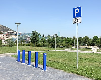 Auf einem grau gepflasterten Weg stehen mehrere blaue Metallbügel, daneben ein Schild mit einem großen weißen P auf blauem Grund und einem Fahrrad-Symbol. Der Parkplatz liegt in einem Park mit großen Rasenflächen.