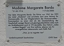 In schwarzer Schrift auf weißem Untergrund ist zu lesen: Madame Margarete Bardo *31.05.1916 +17.04.200 Hier lebte Margarete Bardo. Sie führte von 1961–1991 das Lokal „Madame“ und errichtete dort eine Heimat für Homosexuelle. Sie war Wegbereiterin der Homosexuellen. Emanzipation zu einer Zeit als die Geisteshaltung der Gesellschaft weniger liberal und gleichgeschlechtliche Liebe noch ein Strafbestand nach § 175 darstellte. Das Faktum „Liebe verdient Respekt“ war ihre Leitlinie, ebenso wie ihr unvergeßliches Lebensmotto nach Edith Piaf: Non, je ne regrette rein! Landeshauptstadt Saarbrücken LSVD Saar