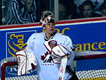 Hockey player in white uniform and goaltender's uniform. He leans against a goal, his helmet lifted above his head, and looks upward.