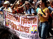 Image of three people walking in a crowd, smiling and holding a banner that reads "LESBIAN HERSTORY ARCHIVES".