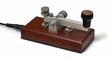 Colour photograph of a telegraph key. A simple metal lever-like switch on a small wooden base connected with a wire disappearing out of frame.