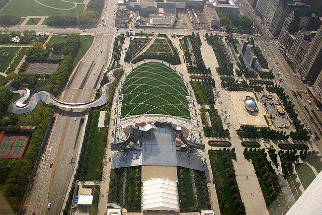 Aerial view of a green park with large roads running vertically at left and right and horizontally at the top. A curving metal bridge crosses the road on the left. Sidewalks divide the park into different areas, and it includes buildings and sculptures.