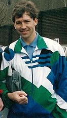A man with brown hair, wearing a white jacket with a pattern of green, blue and black shapes all over it, holding a small grey bag under his right arm and smiling broadly