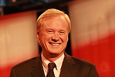 A smiling man with blond hair wearing a gray suit jacket and black tie.