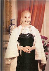 A picture of a Evita, former first lady of Argentina. Her hair is drawn into a tight bun at the back. She is wearing a black, low-cut dress. Around her neck is a number of chains. The lady's hands are folded in her front and she has a white fur shawl around her.