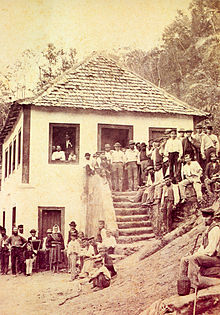 Photograph showing a crowd of people gathered around and on the steps of a white stuccoed house with a hip roof covered in wood shingles situated on a steep slope of a forested hill
