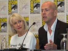 Bruce Willis speaks at San Diego Comic-Con. Actress Helen Mirren is seated to his right wearing a white shirt with the name Harvey Pekar