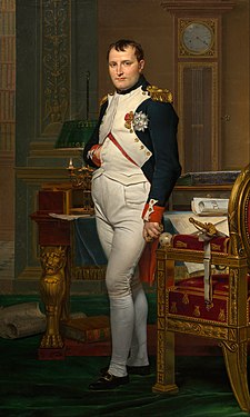 Full length portrait of Napoleon in his forties, in high-ranking white and dark blue military dress uniform. He stands amid rich 18th-century furniture laden with papers, and gazes at the viewer. His hair is Brutus style, cropped close but with a short fringe in front, and his right hand is tucked in his waistcoat.