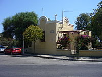 Durrell’s house in Rhodes features Mediterranean architecture and has yellow-painted stucco or plaster walls. It is located on a paved asphalt street, with two cars parked parallel to it. The house is surrounded by several trees, shrubbery, roses, and flowering bushes.