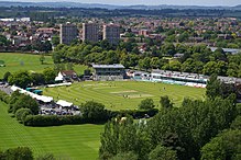 Aerial photo of a cricket ground with trees in the foreground and an urban area behind