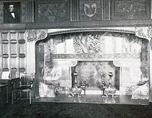 A black and white photograph of a fireplace with ornate mantel set in wood-paneled wall with similarly ornate carvings above it