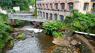 Vue en plongée de l'usine du May. La moitié gauche est occupée par la Durolle traversée dans cette partie par 3 ponts métalliques. La moitié droite présente la façade de l'usine avec 2 étages d'ateliers à 8 vastes fenêtres voutées chacun. Au niveau bas les fenêtres sont voutées de pierres taillées, tandis qu'au niveau haut elles le sont de briques rouges.Le nom de l'usine peint sur la façade est encore bien lisible