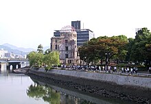 A shot along a river. There is a bridge in the distance, and a ruined domed building in the middle distance. People walk along the footpath that runs parallel to the river.