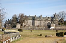 Photo d'un château gris avec des tourelles dont le parc s'étandant à ses pieds rejoint la piste d'un hippodrome.