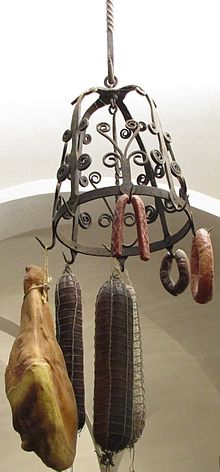 Une couronne d’office en fer forgé formant un dôme ajouré, décoré de volutes, est suspendue par une barre de fer torsadé au plafond en voute blanche du Musée de la Gourmandise; un gros jambon and des saucissons de tailles diverses sont attachés à ses crochets par des ficelles.