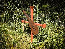 Photographie en couleur d'une croix orange sur lequel est fixé une guitare stylisée dans un matériau qui ressemble à du contreplaqué. La croix est plantée dans un sol recouvert de végétation.