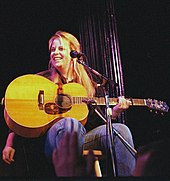 A woman with long blonde hair sitting behind a microphone and holding a guitar