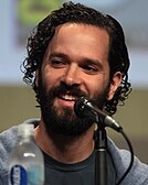 A 35-year-old man with curly black hair talking into a microphone, looking at something to the left of the camera.