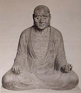 Front view of a cross-legged seated statue with long earlobes. His hands rest on his knees with the palm of his right/left hand turned down/up. Black and white picture.