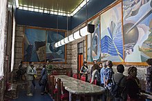 L'intérieur d'une grande salle ; sur deux murs, on voit cinq toiles représentant cinq formes de communication.