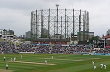 A cricket match in progress; gasholders are visible in the background