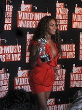 Cette image nous présente une femme brune afro-américaine portant une robe décolleté et courte rouge lors de son passage sur le tapis rouge. Elle a les cheveux bouclés et elle tient de la main gauche un moonman qui est le trophée des MTV Video Music Awards. Le fond est noir avec des logos de la cérémonie.