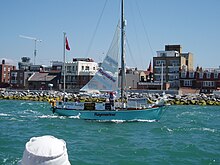 blue-hulled two-masted yacht with mainsail partly set passing a rocky shoreline with buildings beyond