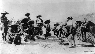 Um grupo de 14 homens armados, 8 dos quais acocorados, com chapéus e segunrando espingardas
