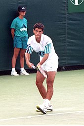 A black-haired man in white shorts and a white shirt prepares to serve with a modern racket