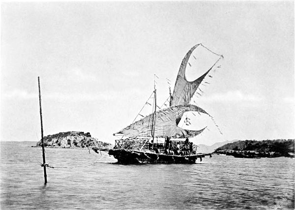 Black and white photograph of a moored sailing boat, with an island visible a short distance behind it.