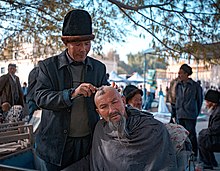 Uyghur man having his head shaved in a bazaar. Shaving of head is now seen mostly among the older generations. Qoy Baziri (198247507).jpeg