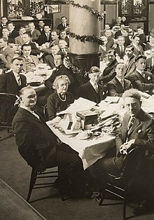 Photo noir et blanc d'un ensemble de personne autour de plusieurs tables sur lesquels se trouvent des cadeaux de Noël déballés. La salle est ornée de guirlandes et autres décoration de Noël.