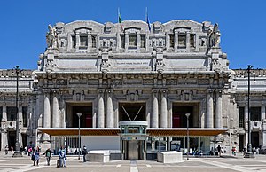 An ornate and extensively detailed gray stone classical portico with four sets of twinned columns at the top of which the Italian and European Union flags hang limply; above is clear blue sky. In front is a more modern one-story pavilion with an elevator in front of it inside a circular wall. Some people can be seen walking around.