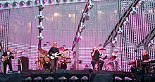 Four men on a stage; two are playing guitars, one is sitting on a stool and holding a microphone, and one is playing keyboards. Various stage equipment, lighting fixtures, drum sets, speakers and other audio equipment can be seen in the background.