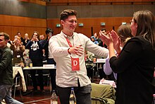 Philipp Türmer (Bildmitte) in gestreiftem Hemd und mit Konferenz-Badge um den Hals geht freudestrahlend und mit offenen Armen auf eine rechts im Bild stehende Person zu.