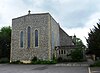 Front view of a flint church with a tall square façade topped with a small stone cross and with two round-headed windows flanking a taller, narrower one. There is a paved parking area in front.