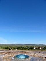 Circle of water in the ground with houses and a green skyline in the distant background.