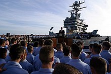 A man in a dark suit and tie speaks to sailors on an aircraft carrier. A sign in the background reads "Mission Accomplished"