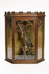 A stand microscope created by Joseph Gutteridge, a weaver from Coventry. The microscope allows coarse and fine adjustments, and its height is adjustable by a set screw. It consists of two objectives each with a screw on cap and a slidecarrier that is moveable laterally. The mirror also has a cap. It has a bench magnifier, lens moveable through 360° and has a base weighted with lead. It is placed in a Mahogany box containing seventeen six-compartment trays of slides.