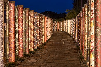 Vista noturna da “Floresta dos Quimonos”, uma coleção de 600 quimonos enrolados em postes com iluminação LED em seu interior, localizado na estação de Arashiyama, distrito de Ukyo, Quioto, Japão. O projeto de decoração foi desenvolvido pelo designer de interiores Yasumichi Morita e consiste em peças de tecido tingidas no estilo Kyo-yuzen, revestidas com fibra acrílica e em forma de postes cilíndricos de dois metros de altura instalados ao redor da estação e dos trilhos do trem. (definição 6 585 × 4 390)