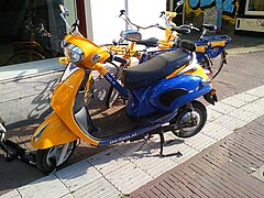 Farbfoto. Blau-gelber E-Roller von Novox im städtischen Umfeld, angeschlossen an Vorderrad. Im Hintergrund weitere Leihfahrräder von OV-fiets sowie ein weiterer Roller.
