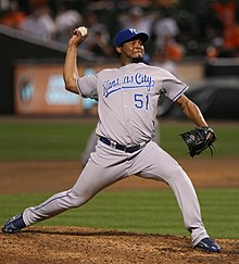 A brown-skinned man with a goatee wearing a gray baseball uniform with blue trim and a blue baseball cap throwing a baseball with his right hand