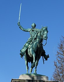 Statue représentant un homme assis sur un cheval, dont la jambe avant droite est levée, tenant un sabre levé dans sa main droite, le tout sur fond de ciel bleu.