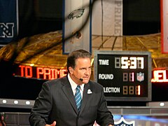 Откровенная фотография Мариуччи, сидящей за столом на телевизоре в темном костюме в тонкую полоску, галстуке в синюю полоску и наушниках.