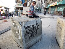 A Palestinian child sitting on a roadblock at Al-Shuhada Street within the Old City of Hebron in the Israeli-occupied West Bank. Palestinians have nicknamed the street "Apartheid Street" because it is closed to Palestinian traffic and open only to Israeli settlers and tourists. Austin 202 (8469717987).jpg