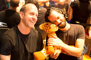 Мужчина с короткими каштановыми волосами, сидящий рядом с мужчиной с вьющимися черными волосами, обнимающий плюшевого жирафа, оба улыбаются чему-то справа от камеры.