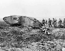Files of soldiers with rifles slung follow close behind a tank, there is a dead body in the foreground